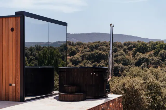 Jacuzzi nórdico de madera con chimenea en terraza exterior con vistas al bosque y montañas.