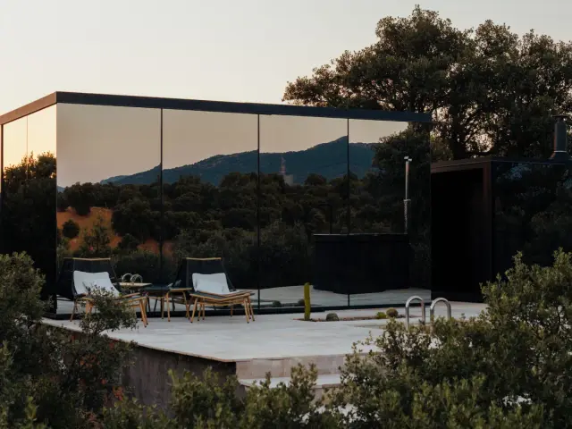 Terraza exterior de casa moderna con espejos, dos tumbonas de madera y paisaje de montañas al atardecer.