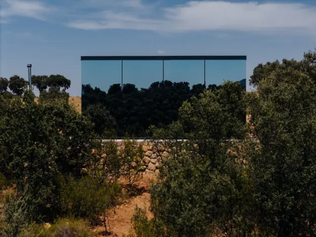 Fachada de casa moderna con fachada de vidrio reflejando árboles, rodeada de vegetación y muro de piedra bajo