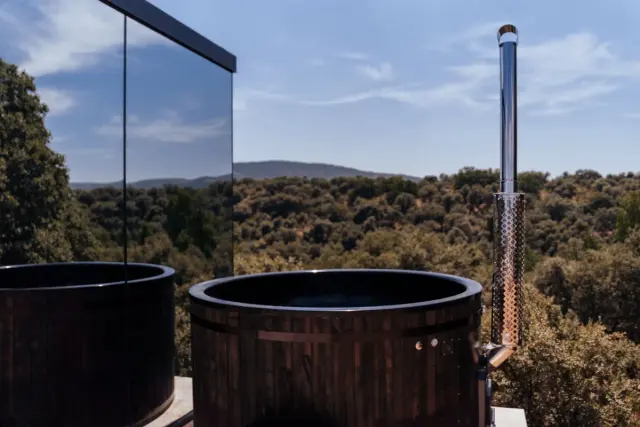 Jacuzzi nórdico de madera con chimenea en terraza frente a un paisaje boscoso.