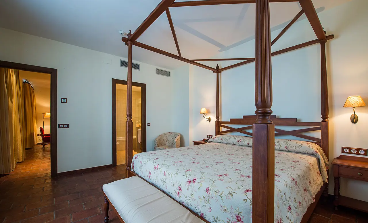 Vista de una suite con una gran cama de dosel de madera oscura, cubierta con colcha floral.