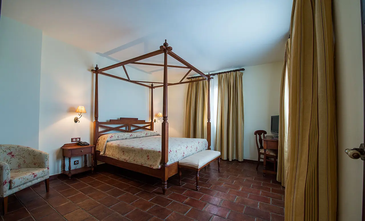 Vista de una suite con cama de dosel de madera, suelo de terracota, butaca floral, escritorio con silla, cortinas