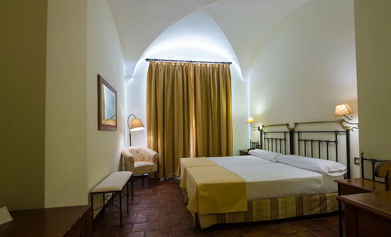 Habitación de suite con cama doble de matrimonio, cabecero de forja, colcha amarilla, cortinas ocres, butaca