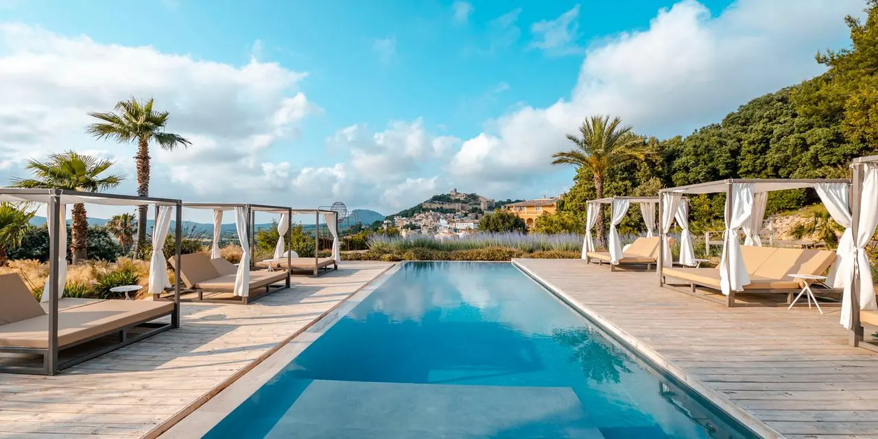 Vista de una piscina rectangular de agua azul rodeada de una terraza de madera con tumbonas y cabanas con cortinas