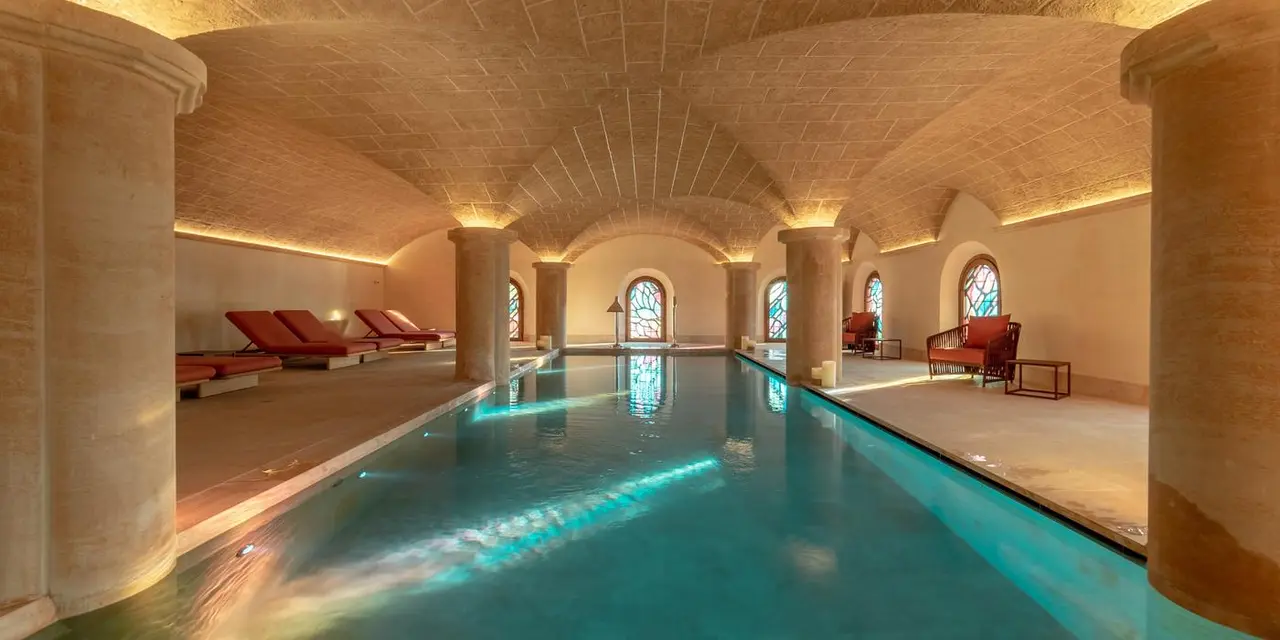 Vista interior de una piscina rectangular iluminada con agua turquesa, rodeada de columnas de piedra y arcos
