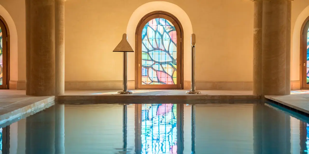 Interior de una piscina rectangular con agua tranquila que refleja una gran ventana arqueada de vitrales de colores.