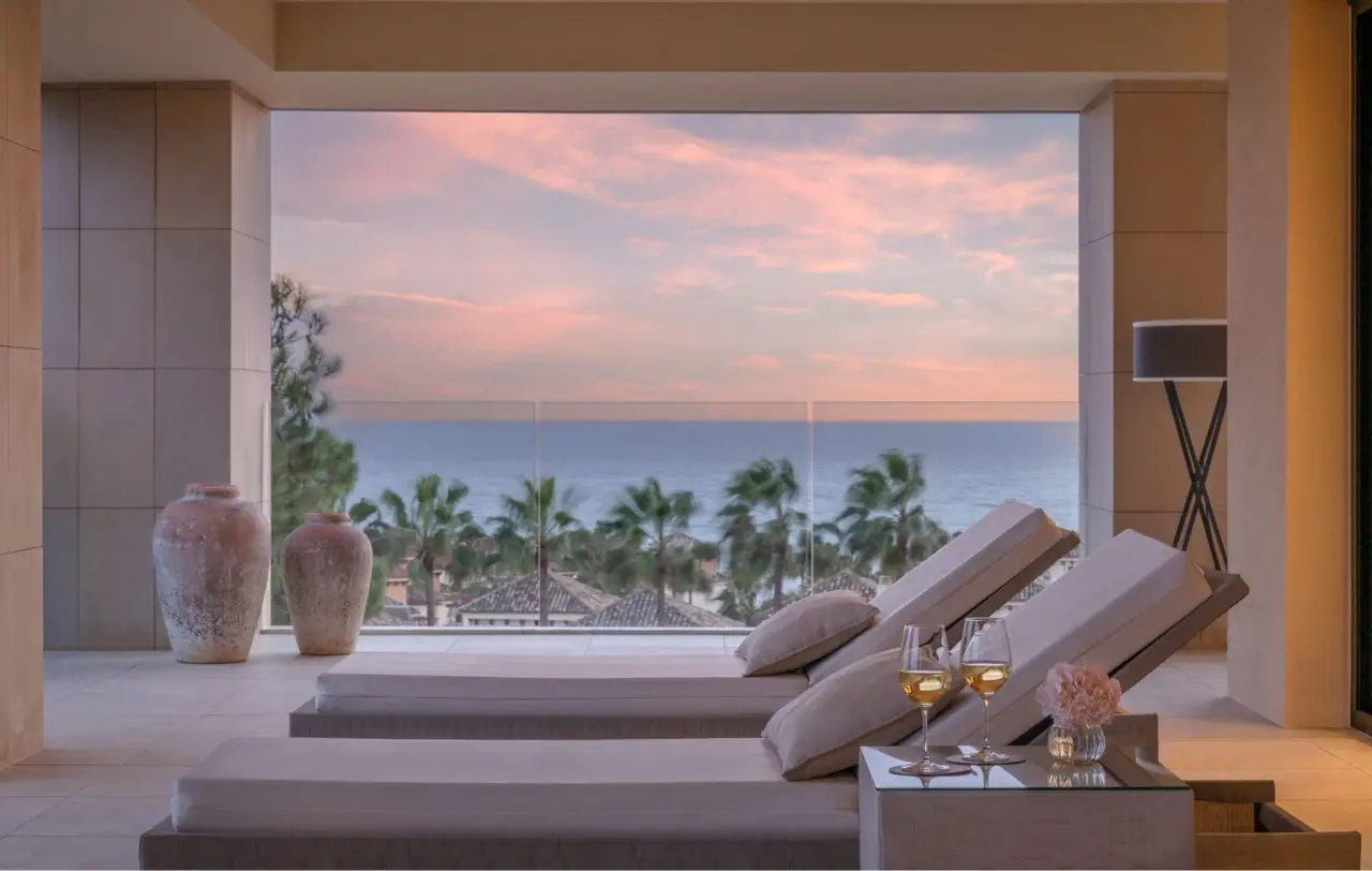 Vista desde una terraza interior con dos tumbonas acolchadas en primer plano, mesa con copas de vino y flores