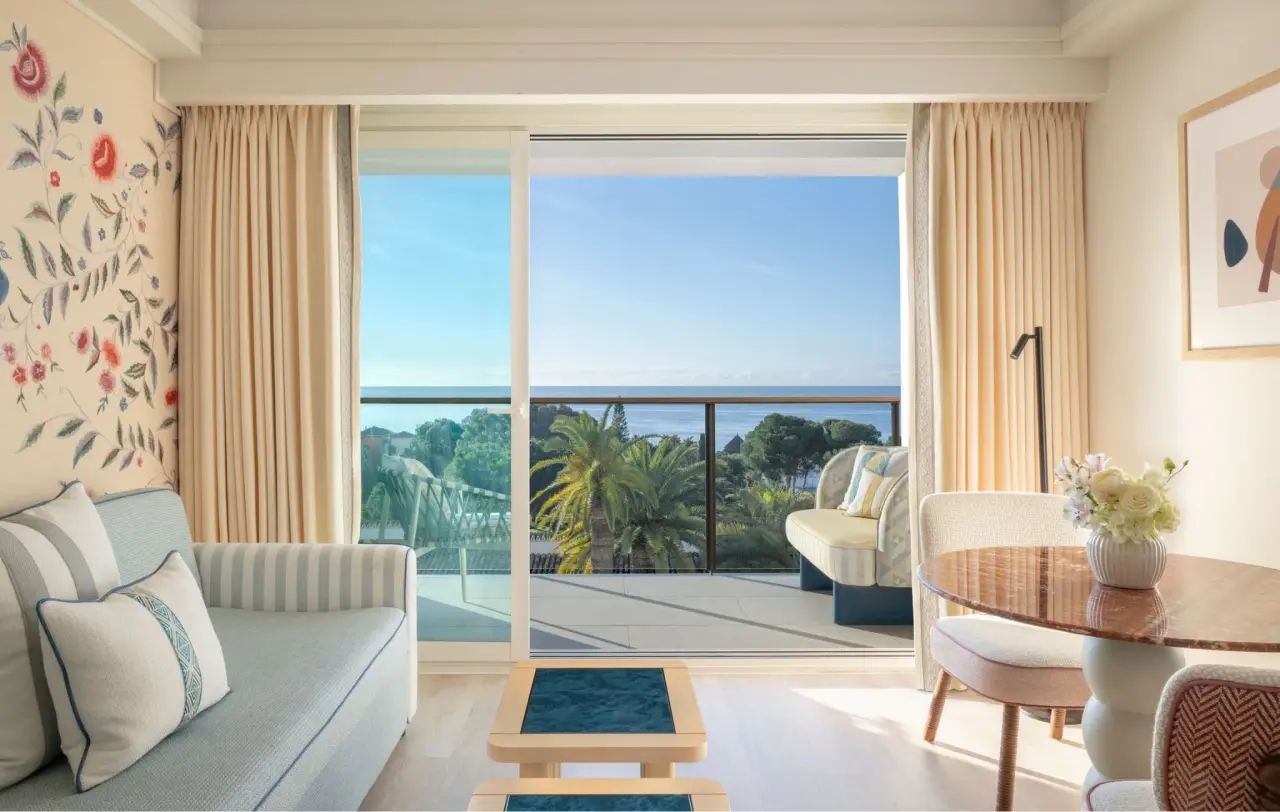 Vista desde el interior de una suite hacia una terraza con barandilla, palmeras y el mar al fondo en un día soleado.