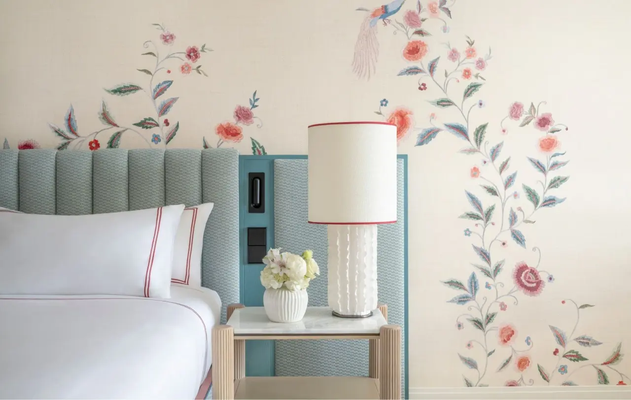Vista de una habitación con cama blanca, cabecero azul texturizado, mesita de noche con lámpara y jarrón de flores