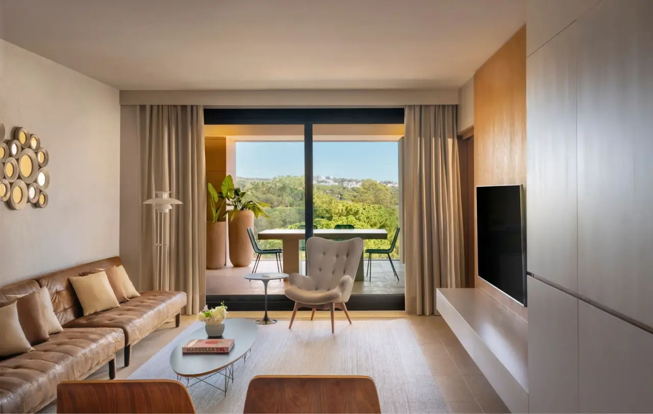 Interior de una suite con sofá de cuero marrón, sillón tapizado, mesa de centro, televisión en la pared y grandes