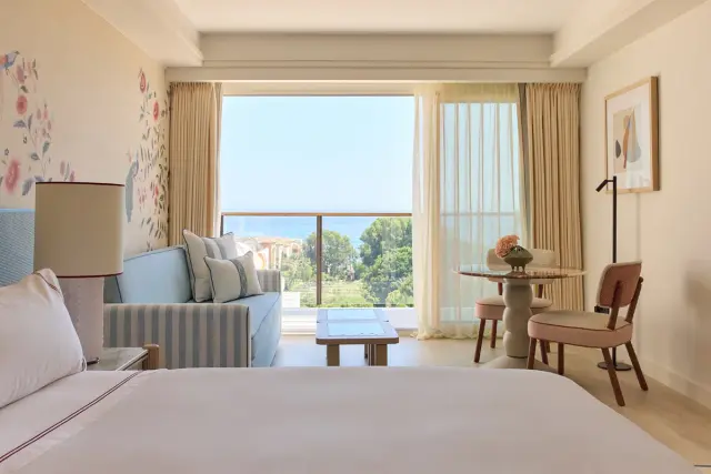 Vista interior de una suite con cama grande en primer plano, sofá azul, mesa de centro, zona de comedor con mesa