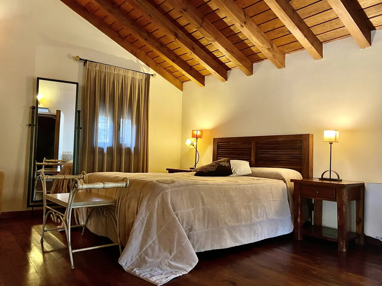 Interior de una habitación con cama doble, cabecero de madera, mesitas de noche con lámparas encendidas, silla