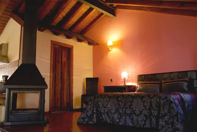 Interior de una habitación con cama doble, chimenea de leña, puerta de madera, silla y lámparas de pared y mesa.
