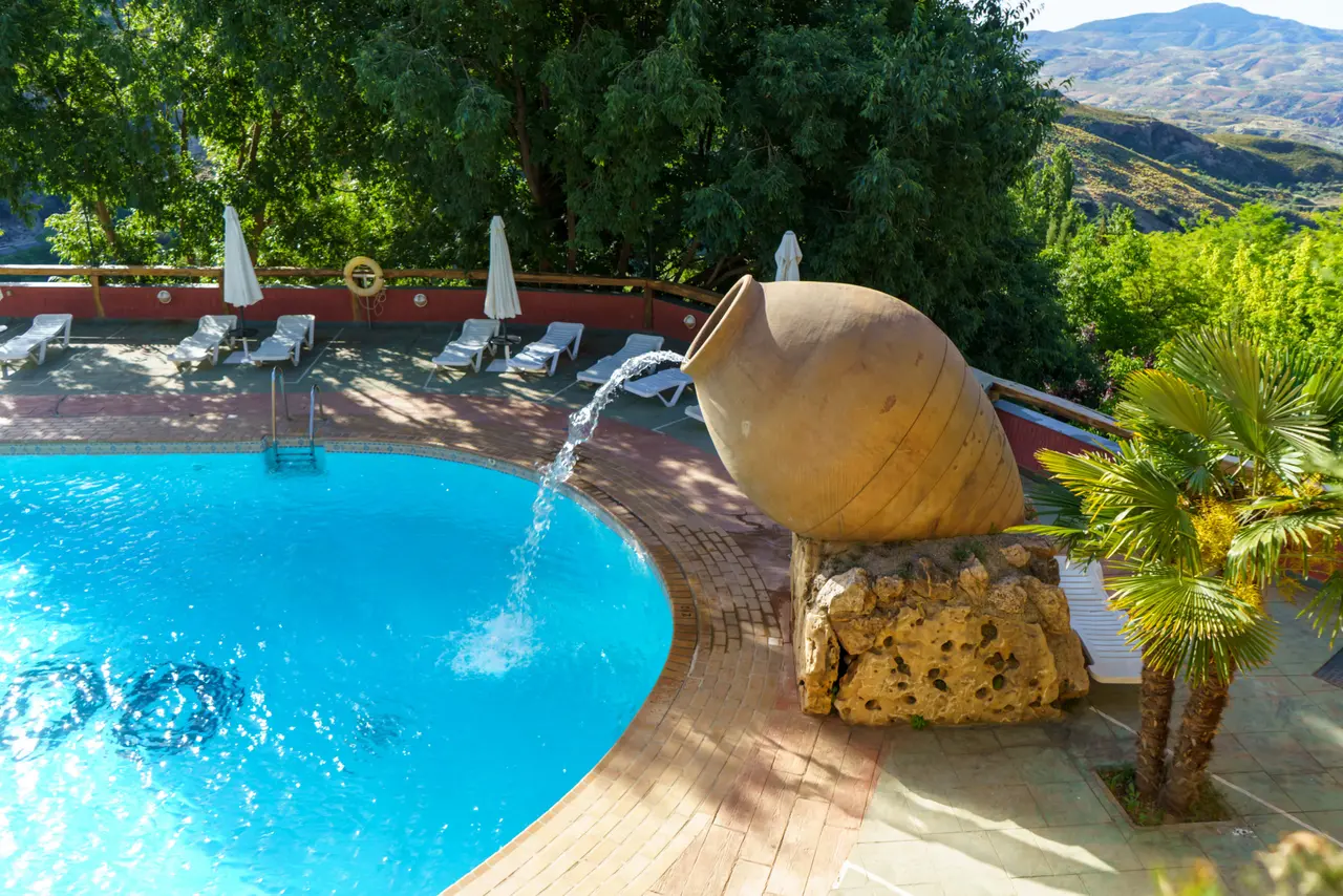 Piscina exterior con agua azul, rodeada de tumbonas, sombrillas y un gran jarrón de terracota que vierte agua.
