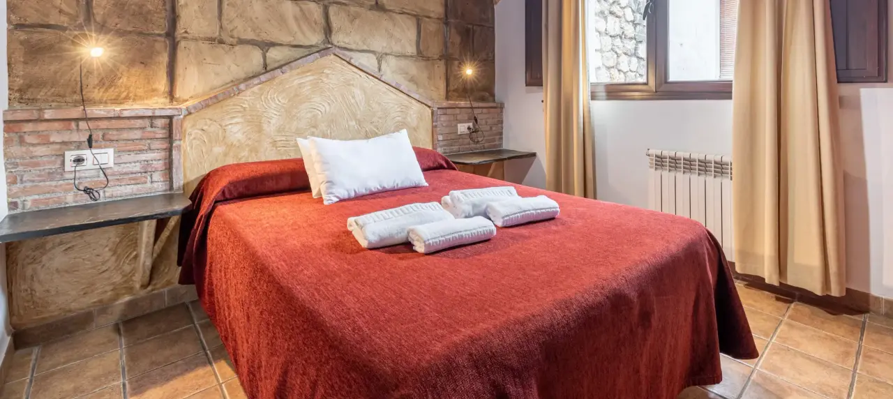 Habitación con cama doble cubierta por una colcha roja, cabecero de piedra y ladrillo visto, dos mesitas de noche