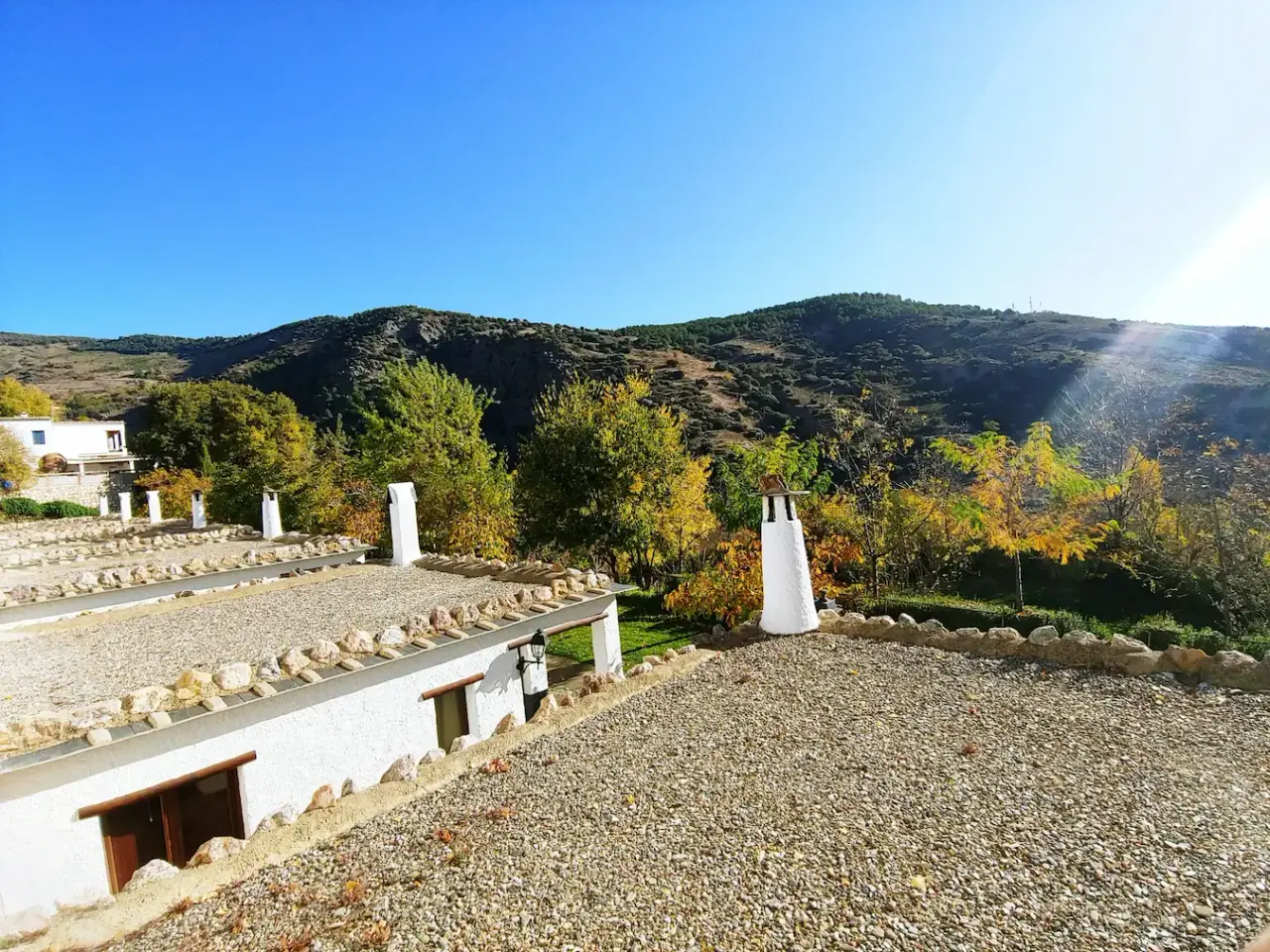Vista exterior de un alojamiento rural con techos de grava y chimeneas blancas, rodeado de vegetación otoñal