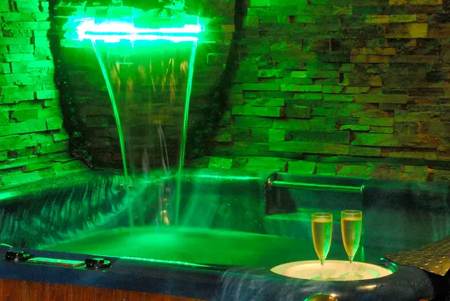 Jacuzzi iluminado con cascada de agua verde y dos copas de champán en el borde.