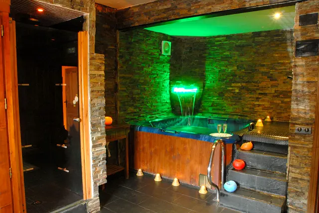 Spa privado con jacuzzi iluminado en verde, cascada de agua y paredes de piedra en una sala con acceso desde un pasillo.