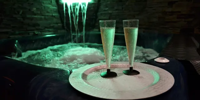 Dos copas de cava sobre una bandeja en el borde de un jacuzzi iluminado con cascada de agua en un spa privado.