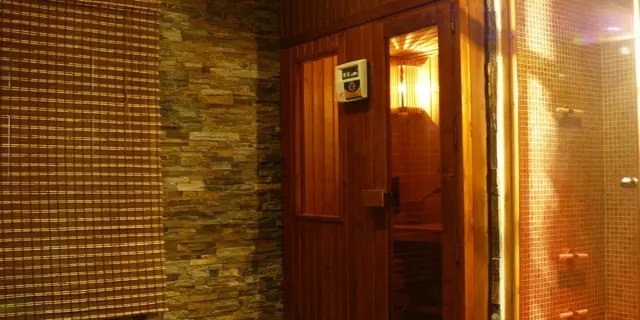 Puerta de madera abierta a una sauna iluminada, junto a una ducha con mosaicos y pared de piedra.