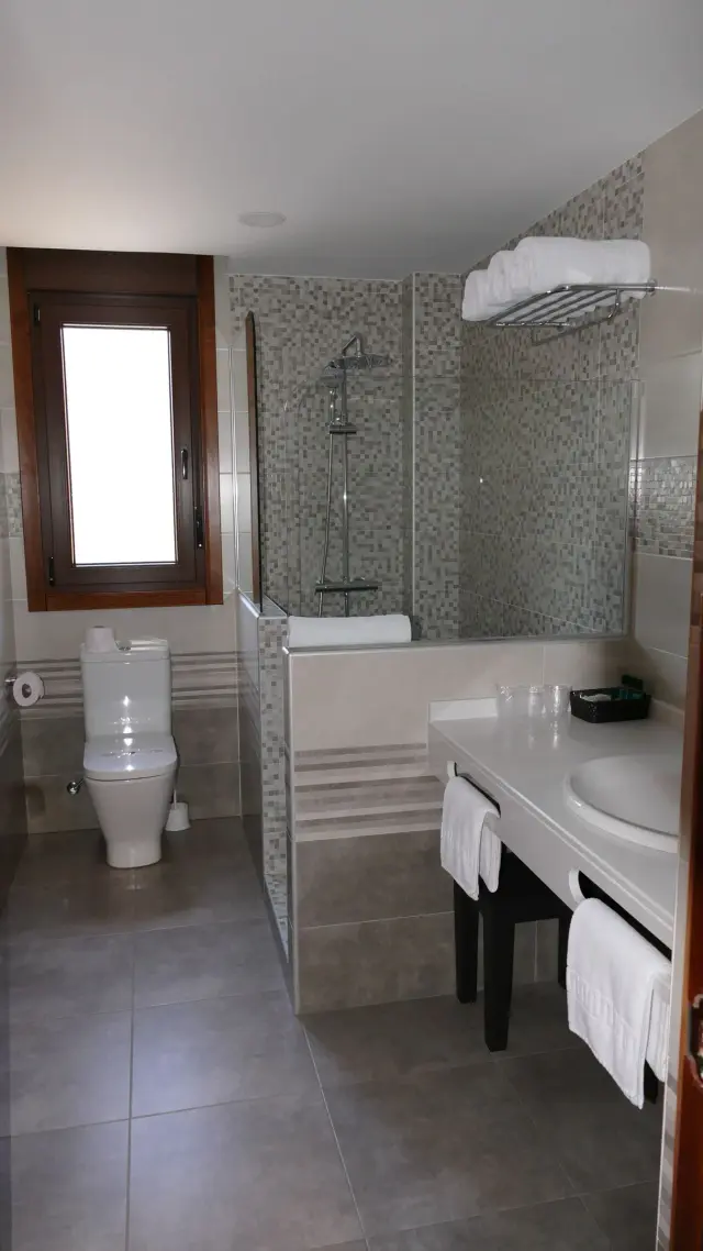 Baño completo con ducha, bañera, inodoro, lavabo sobre encimera blanca con grifo, toallas blancas colocadas en estante