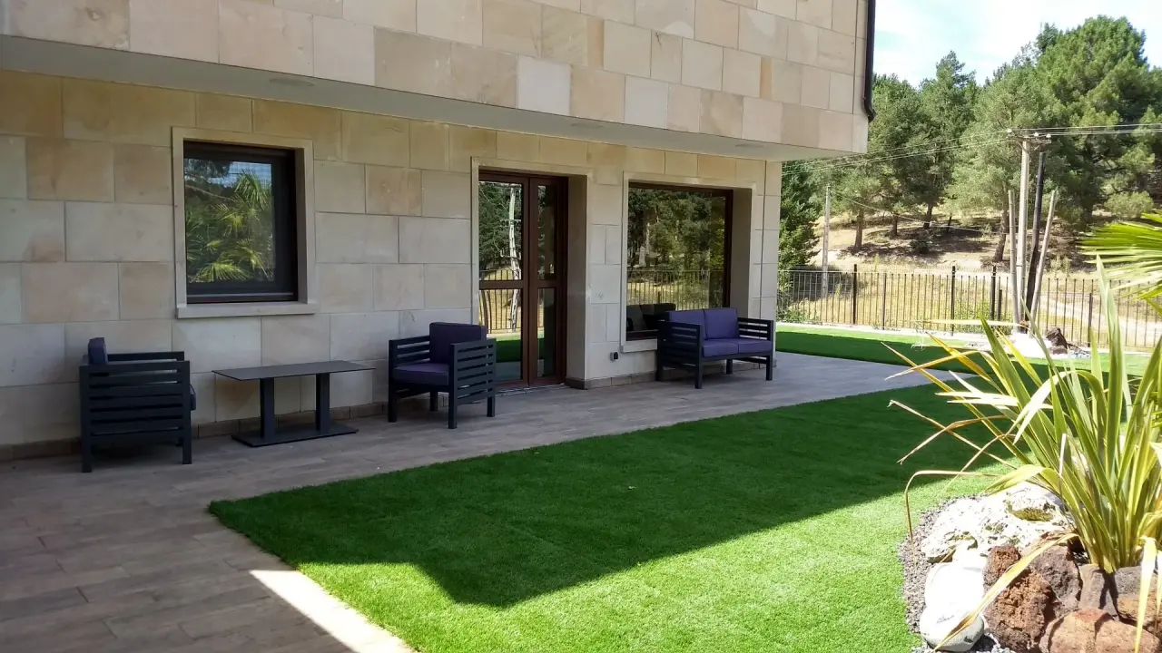 Terraza exterior de una casa con suelo de césped artificial, mobiliario de exterior compuesto por sillones y mesa