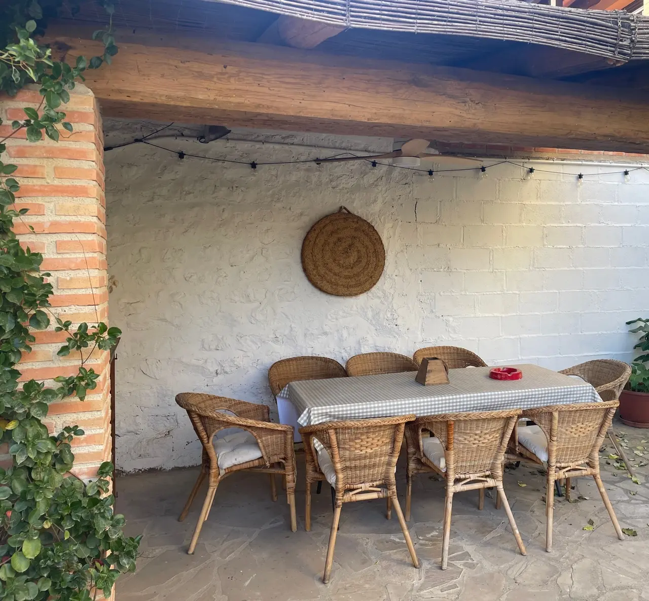 Terraza exterior con mesa rectangular para seis personas, sillas de mimbre con cojines, mantel de cuadros, pared