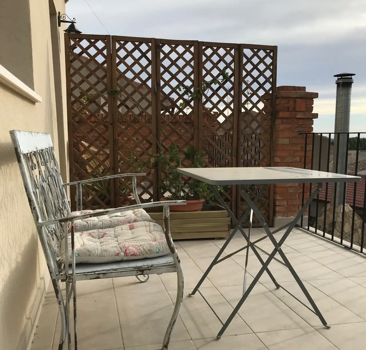 Terraza privada con suelo de baldosas, silla de metal con cojín, mesa plegable, celosía de madera, plantas en macetero