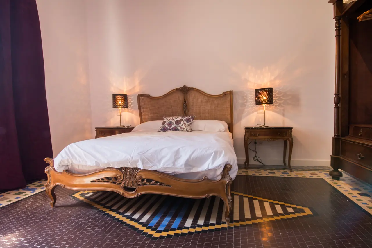 Habitación con cama doble de estilo clásico con cabecero tapizado, dos mesitas de noche con lámparas encendidas, suelo