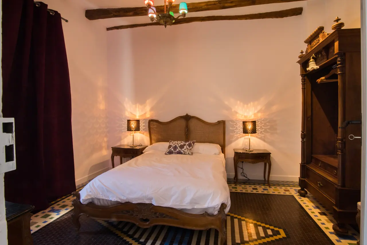 Habitación con cama doble de madera tallada, dos mesitas de noche con lámparas encendidas, armario antiguo de madera