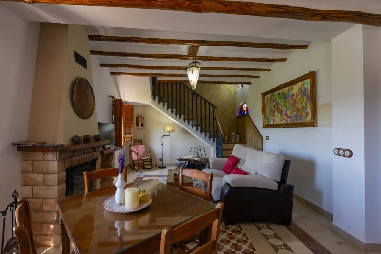 Salón comedor con chimenea de piedra, mesa de madera, sofá y escalera al fondo bajo vigas de madera.