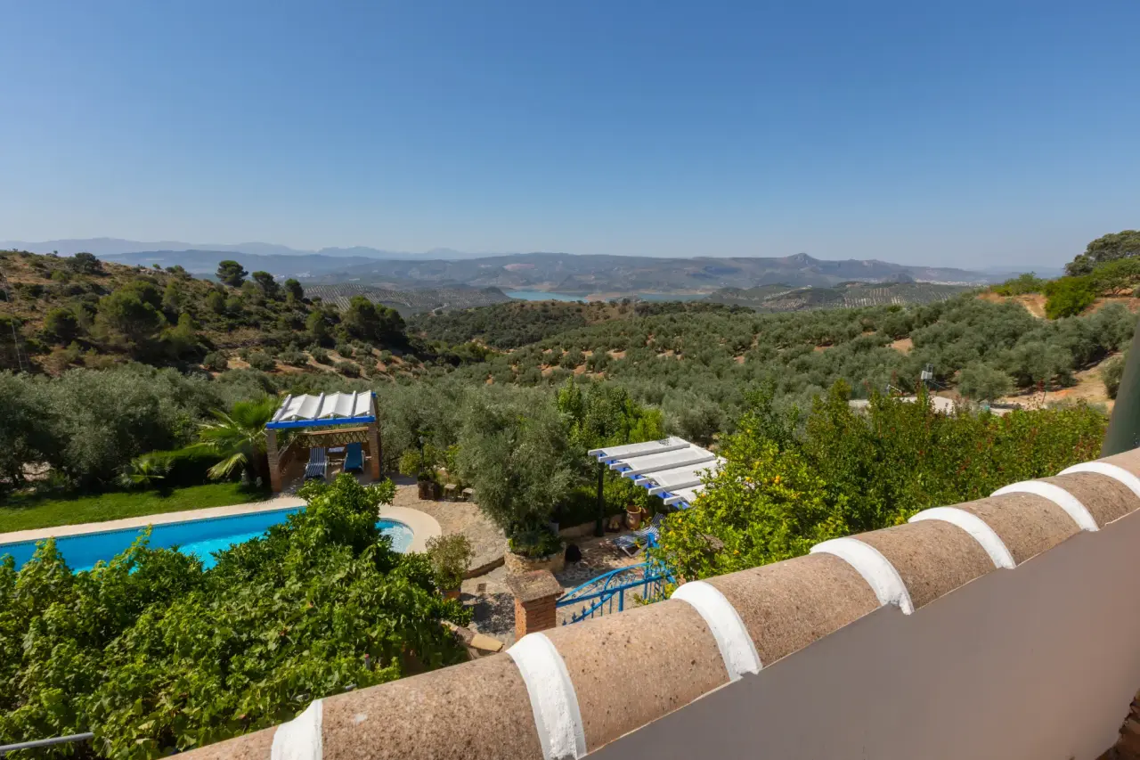 Vista panorámica desde una terraza con barandilla sobre un paisaje de olivos, piscina y montañas lejanas.