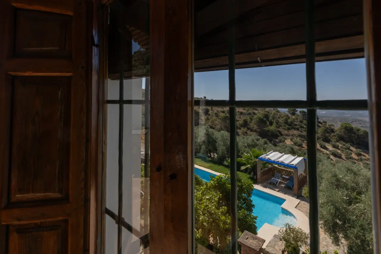 Vista desde una ventana de madera hacia una piscina rectangular en un jardín con olivos y paisaje rural al fondo.