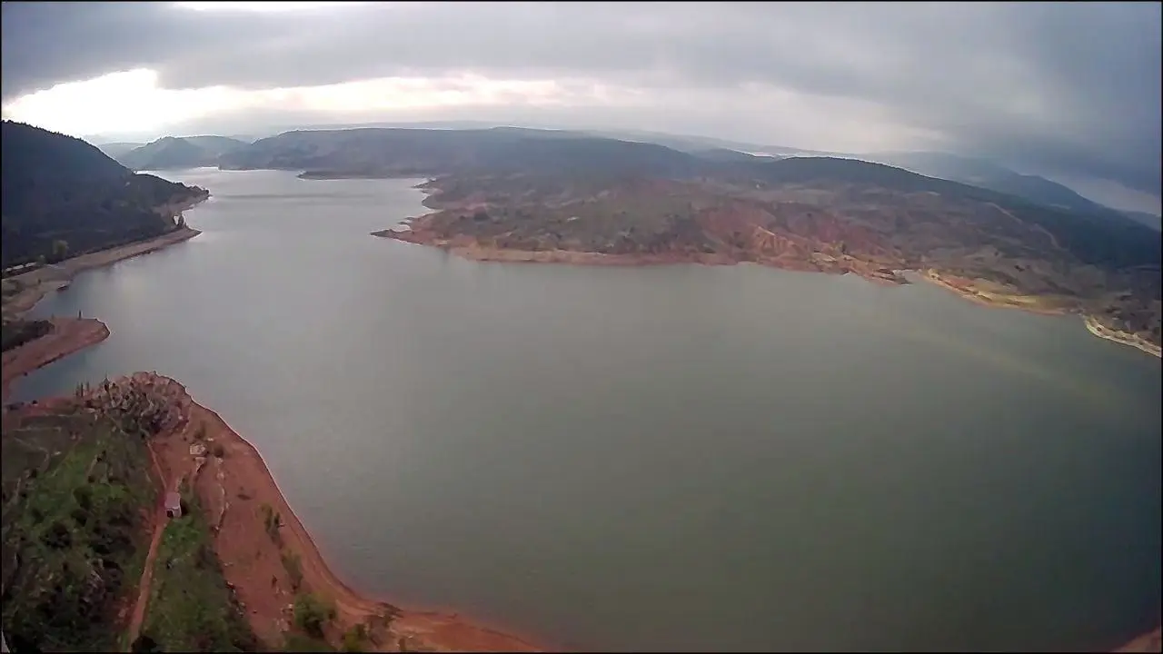 Vista aérea de un embalse rodeado de colinas y montañas bajo un cielo nublado, con orillas de tierra rojiza