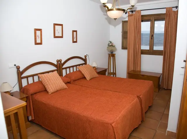 Habitación con dos camas individuales unidas, colchas y cojines naranjas, cabeceros de madera, mesitas de noche