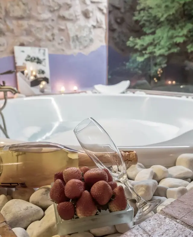 Vista de un jacuzzi blanco en una habitación con paredes de piedra, acompañado de una botella de cava, una copa