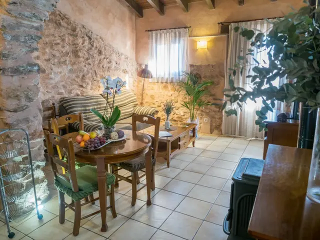 Interior de una habitación rústica con paredes de piedra vista, suelo de baldosas blancas, mesa redonda de madera