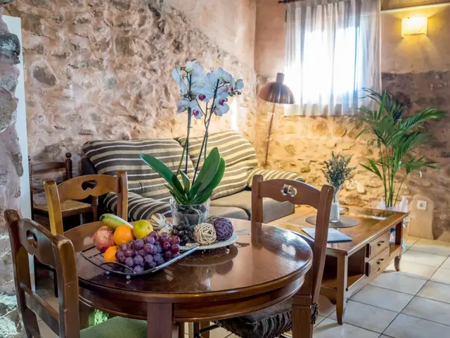 Interior de una habitación rústica con paredes de piedra, mesa redonda de madera con sillas, sofá a rayas, mesa
