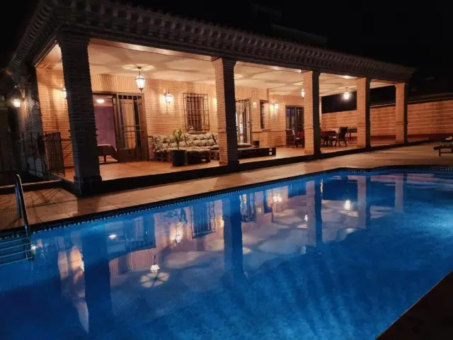 Vista nocturna de una piscina rectangular iluminada con agua azul, frente a una casa rural con porche cubierto