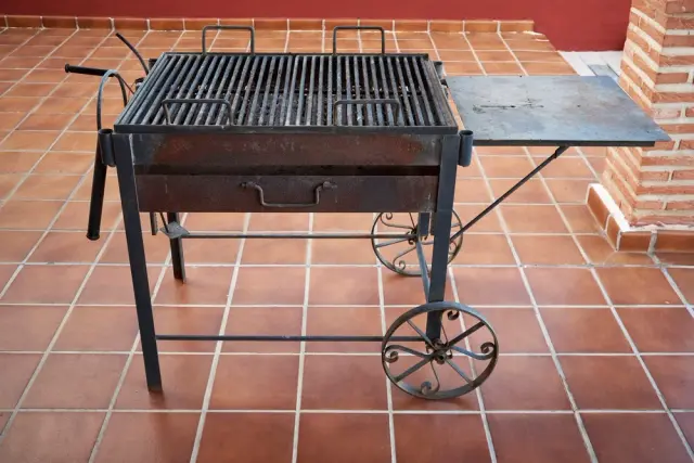 Barbacoa de hierro forjado con ruedas y parrilla rectangular, colocada sobre un suelo de baldosas terracota
