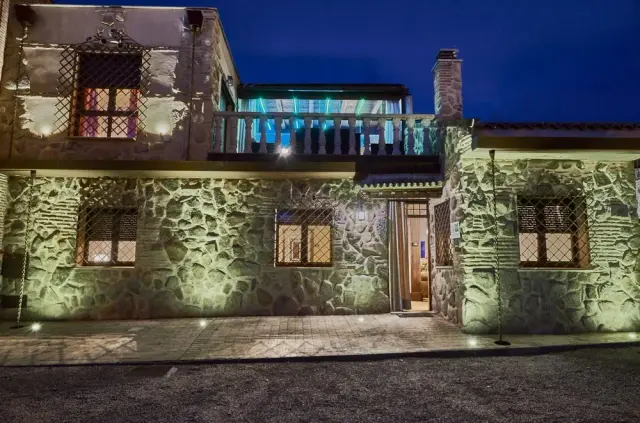 Fachada nocturna de una casa rural de piedra con iluminación exterior, balcón superior con luces azules y patio