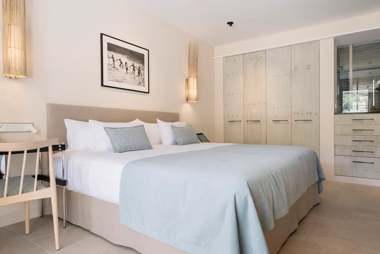 Habitación con cama doble grande vestida en tonos blancos y azul claro, cabecero beige, mesitas de noche, silla