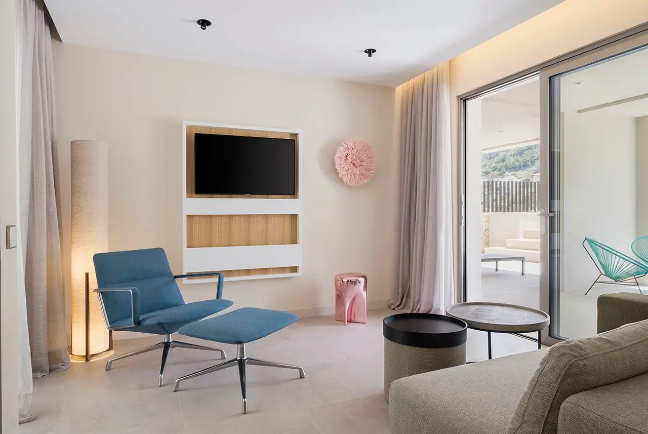 Interior de una suite con paredes claras, suelo de baldosa, un sillón azul con otomana, mesa de centro, televisor