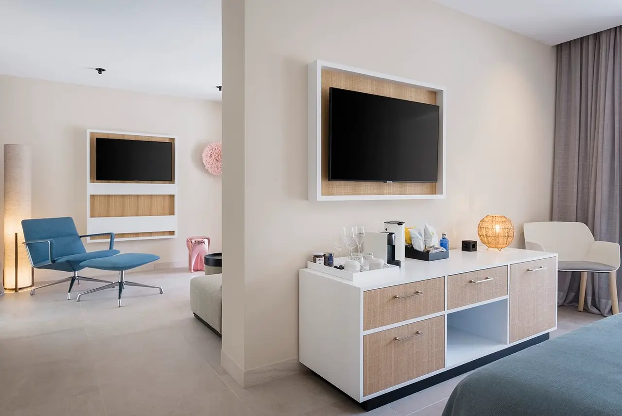 Interior de una suite con zona de estar moderna. Se ve un mueble blanco con TV montada en la pared, una máquina