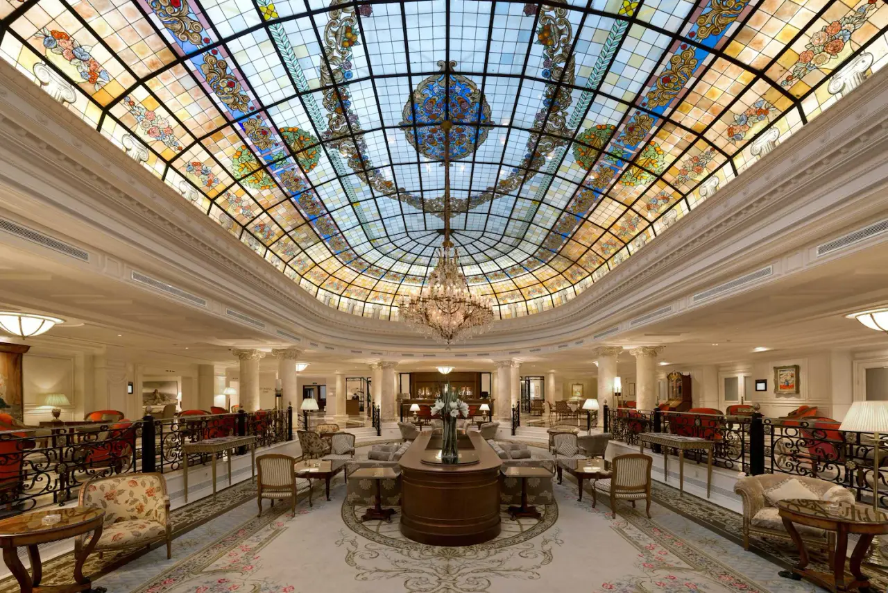 Salón elegante con techo acristalado de vidrieras multicolores y gran araña de luces, rodeado de sofás y mesas