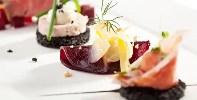 Plato gourmet con base de pan negro, remolacha, jamón ibérico, queso y hierbas frescas sobre superficie blanca.