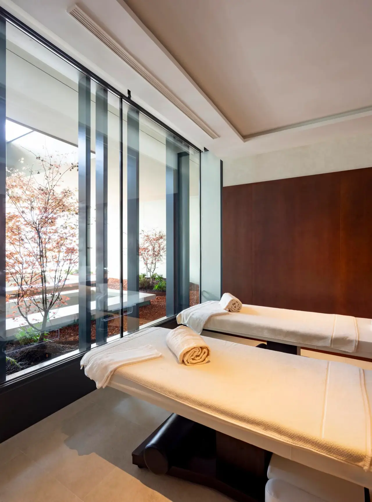 Dos camillas de masaje con toallas enrolladas en una sala de spa con grandes ventanales al jardín.