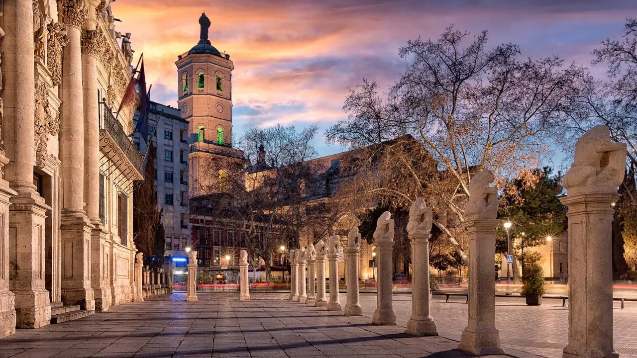Plaza al atardecer con columnas de piedra, estatuas y torre histórica al fondo.