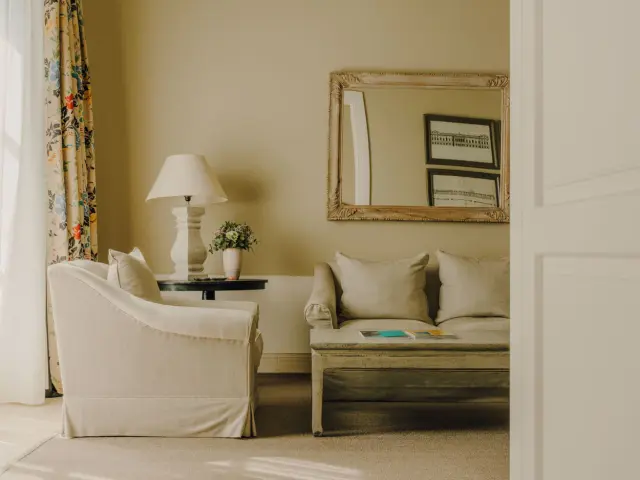 Interior de una sala de estar con un sofá blanco, un sillón, una mesa auxiliar con lámpara y flores, y un espejo