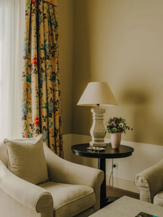 Interior de una habitación con un sillón beige, mesa auxiliar redonda con lámpara y jarrón de flores, cortinas